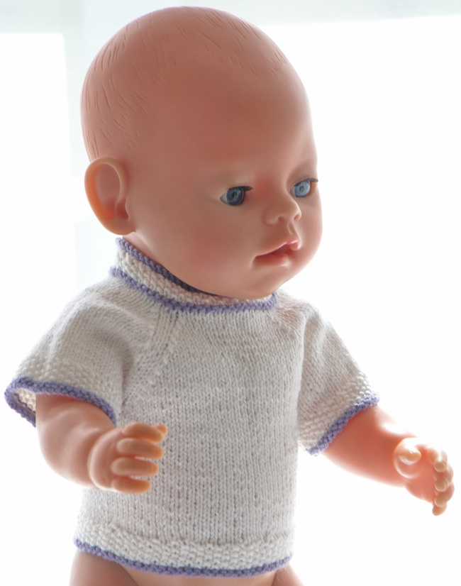 Baby born Kara har fått en enkel, hvit genser. Den er kort-ermet. Kantene er strikket i perlestrikk med enkle kanter strikket i lilla.