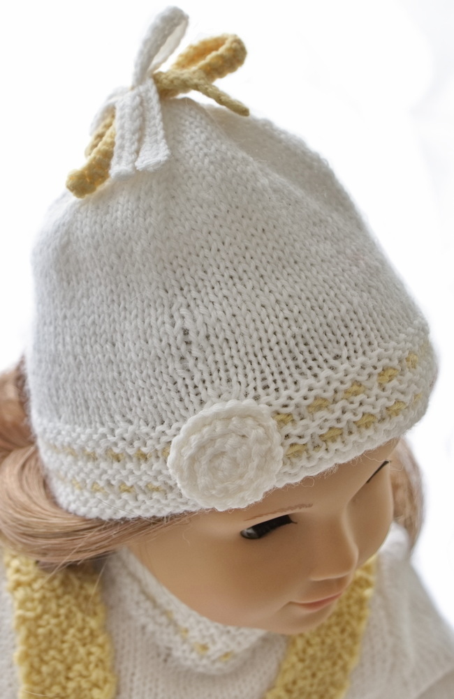 Eine weiße Mütze mit dem gleichen Muster, das auch für den Pullover gestrickt wurde, passt perfekt zu dem Outfit.  Die Mütze ist mit gehäkelten Schleifen versehen, die oben aufgenäht sind.