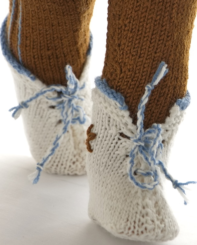 Sehr feine Schuhe in weiß mit einer Kante um die Knöchel, die in blau gestrickt ist. Eine Kordel wird aus einem blauen und einem weißen Faden hergestellt und durch die Löcher gezogen, die an jeder Seite der vorderen Öffnung gestrickt sind. Eine braune Blume ist in Schlingenstichen auf die Außenseite der Schuhe genäht.